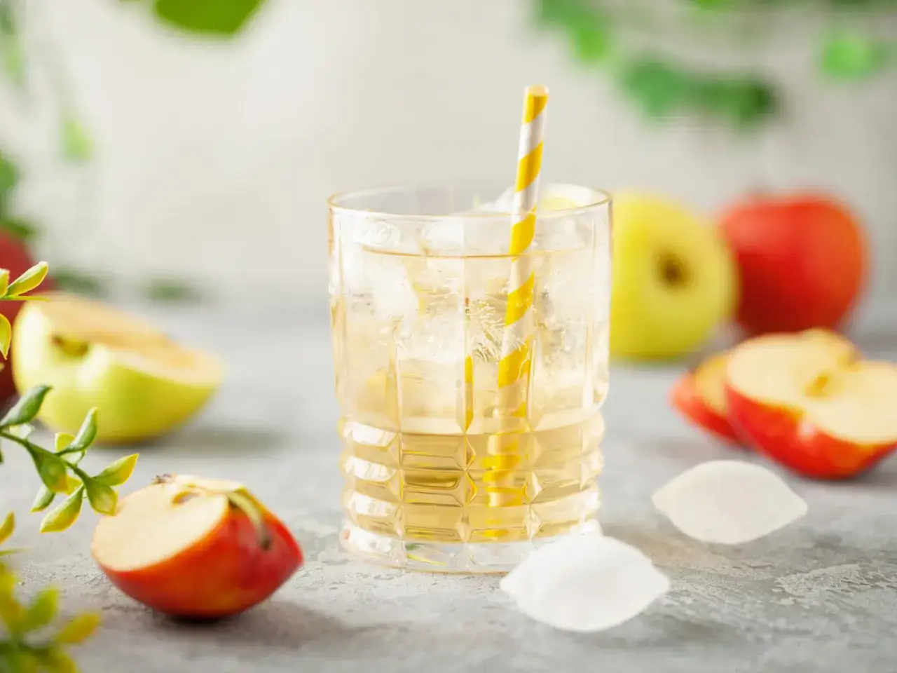 Erfrischendes Apfelwasser selber machen: Ein Glas mit Eis, gelbem Strohhalm und &Auml;pfeln im Hintergrund.