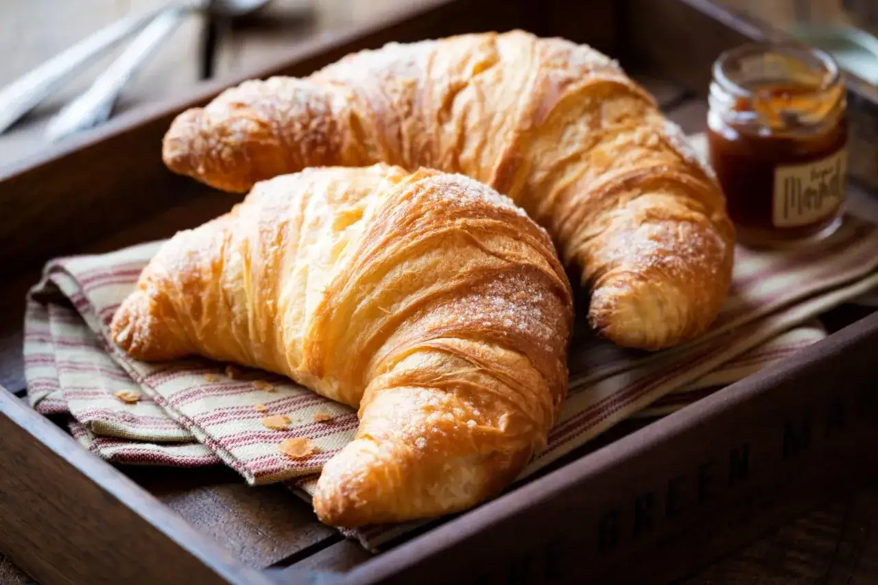 Dwa złociste croissanty posypane cukrem pudrem, obok słoiczek dżemu. Idealne na śniadanie, choć croissant kcal warto mieć na uwadze.
