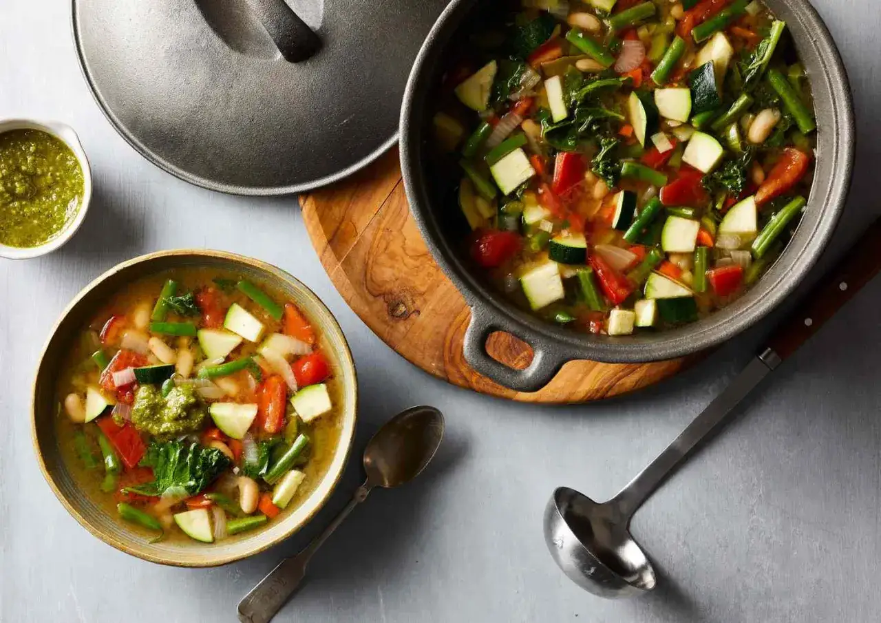 En skål med nyttig soppa gå ner i vikt, full av grönsaker som zucchini, bönor och tomater. En gryta med samma soppa står bredvid.