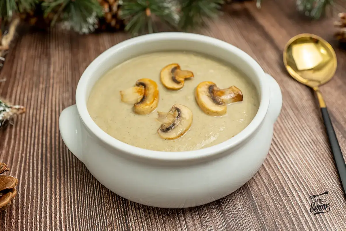 Crema de champiñones servida en un cuenco blanco, decorada con rodajas de champiñones salteados. Una cuchara dorada y ramas de pino completan la escena.