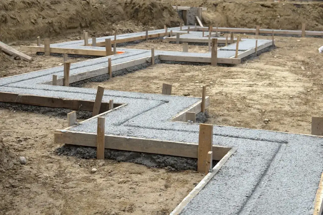 Zbrojenie fundamentów w kształcie litery L, gotowe do zalania betonem. Widać drewniany szalunek i pionowe podpory, tworzące przyszły oczep.