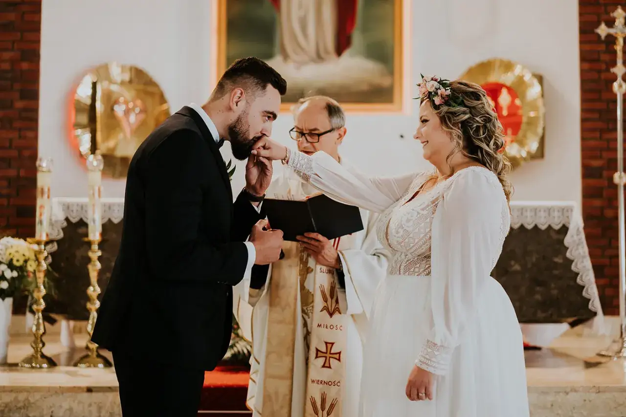 Ślub kościelny i cywilny: pan młody całuje dłoń panny młodej, ksiądz błogosławi parę.