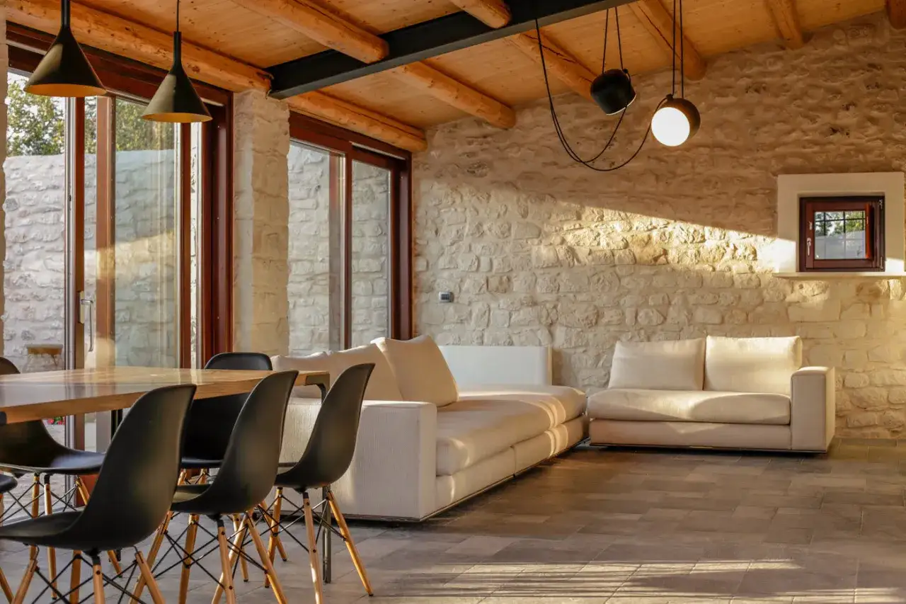 Interni di case di campagna con pareti in pietra, soffitto in legno e arredi moderni. Un tavolo da pranzo con sedie nere e un divano chiaro creano un'atmosfera accogliente.