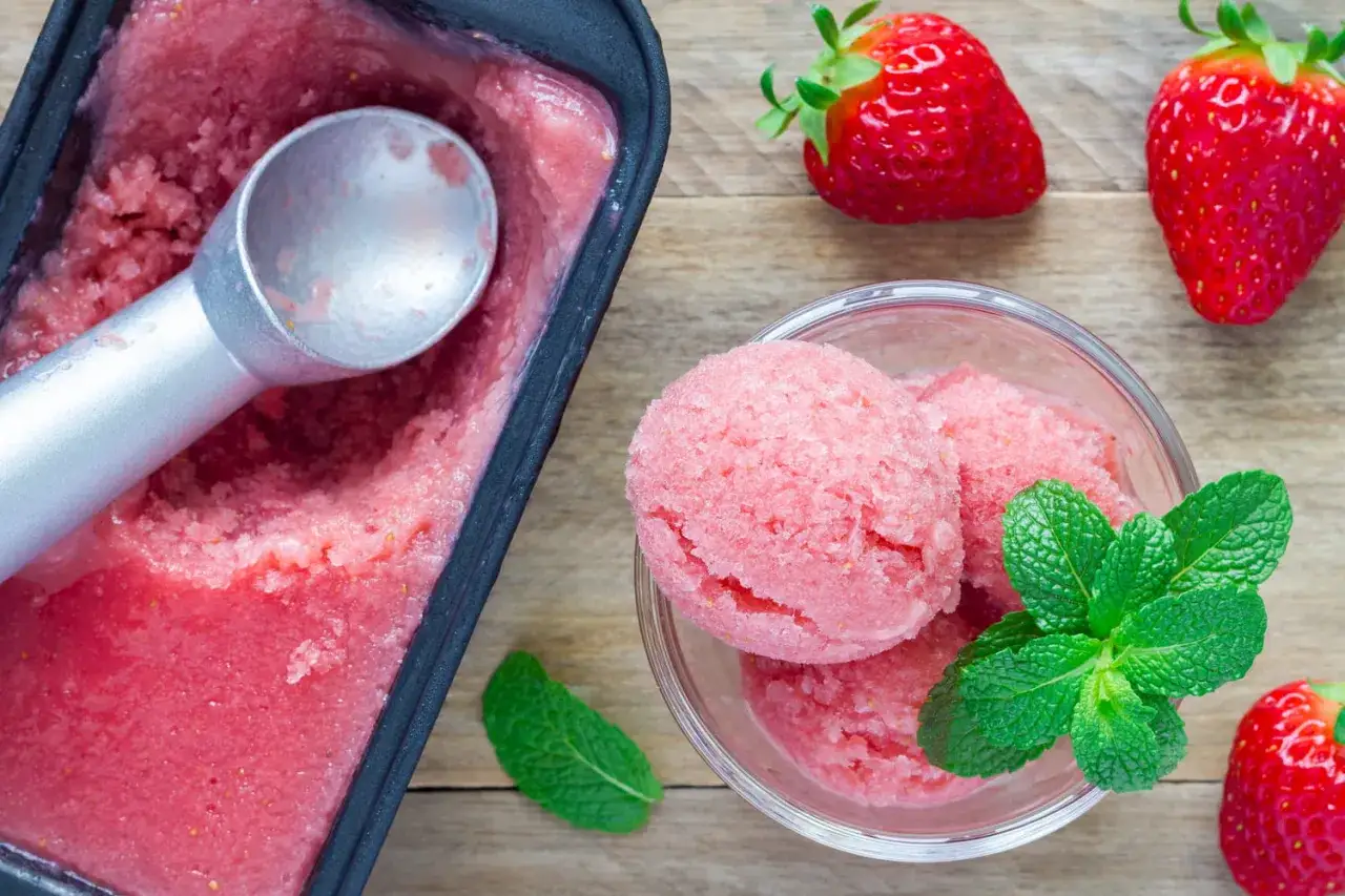 Un délicieux sorbet aux fraises, parfait pour une recette estivale. Des boules roses dans un bol, un bac rempli et des fraises fraîches.