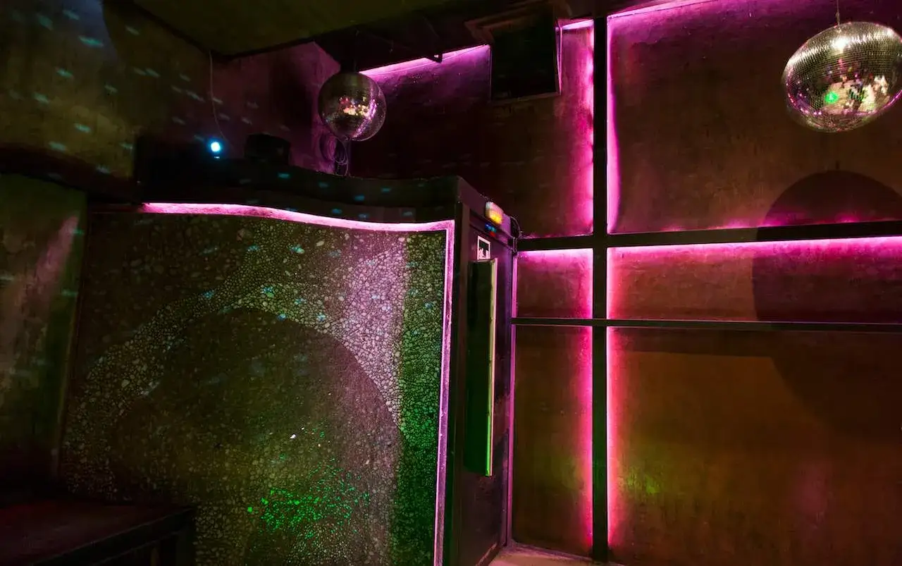 Luci al neon rosa e verdi illuminano un interno di club, con sfere specchiate che riflettono la festa al Macarena Club Barcelona.