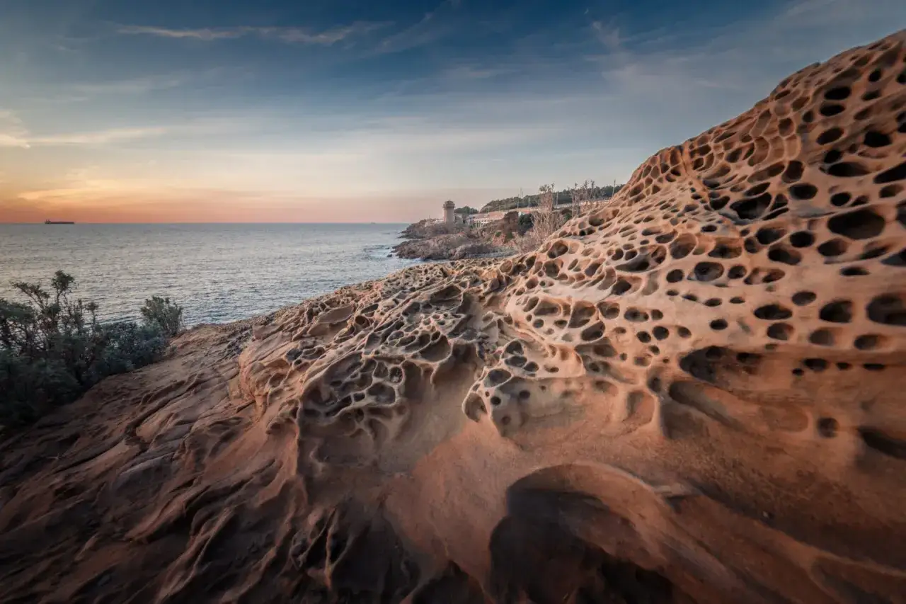 Rocce erose dal mare precisamente a Calafuria, con un faro in lontananza al tramonto.