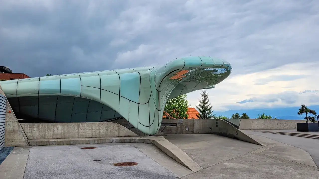 Futuristisches Geb&auml;ude mit geschwungenen Glasfl&auml;chen, ein typisches Beispiel f&uuml;r Zaha Hadid Bauwerke, ragt vor einer dramatischen Wolkenkulisse auf.