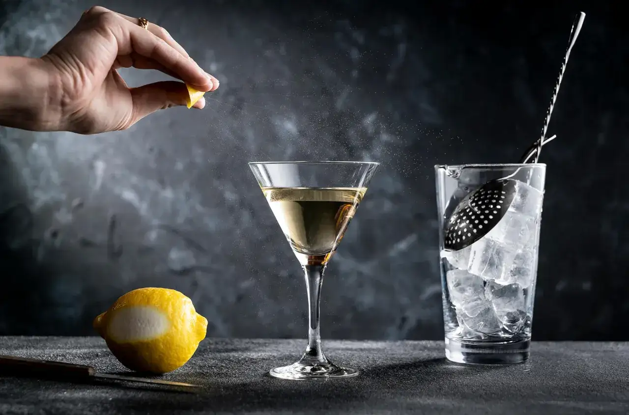En hand pressar en citron &ouml;ver ett martiniglas, som om den f&ouml;rbereder en klassisk James Bond-drink. En citronskal och isbeh&aring;llare syns bredvid.