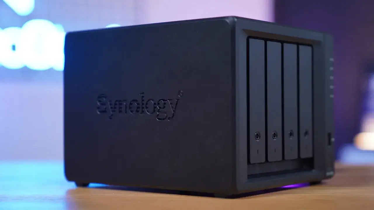 Czarny serwer domowy Synology z czterema zatokami na dyski, umieszczony na drewnianym blacie, oświetlony niebieskim i fioletowym światłem.