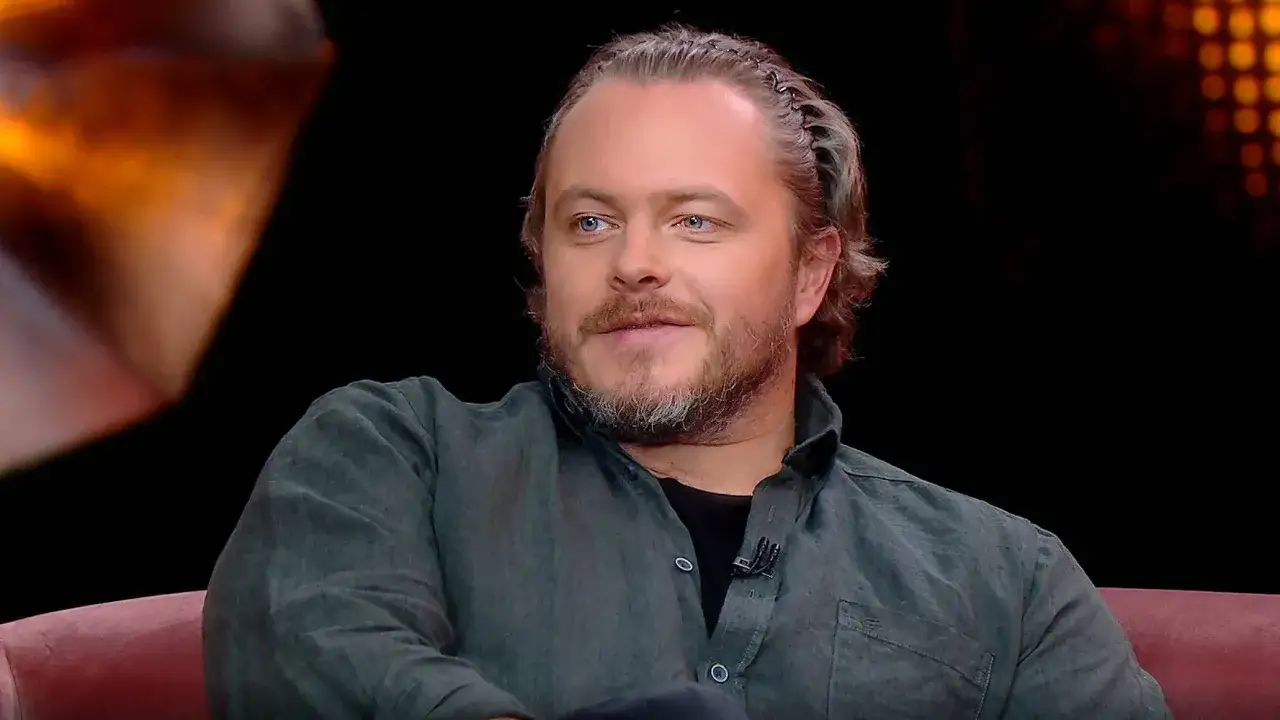 Tomasz Schuchardt, znany z filmów, seriali i programów, z charakterystyczną fryzurą i niebieskimi oczami, siedzi na kanapie.