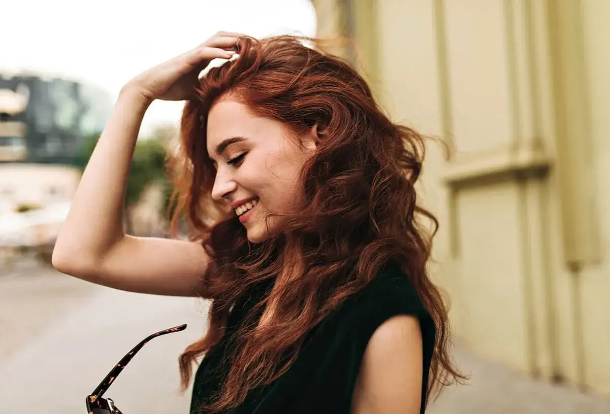 Jeune femme aux cheveux roux, souriant, sa main dans ses cheveux. Peut-être trop d'oxydant dans la coloration a donné ce résultat éclatant.