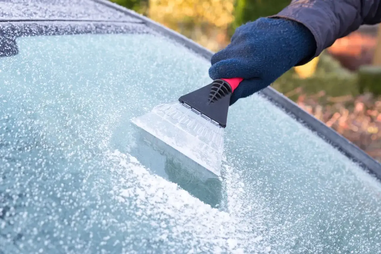 En hand med bl&aring; vante skrapar is fr&aring;n en bilruta. Frosten gnistrar i solljuset.