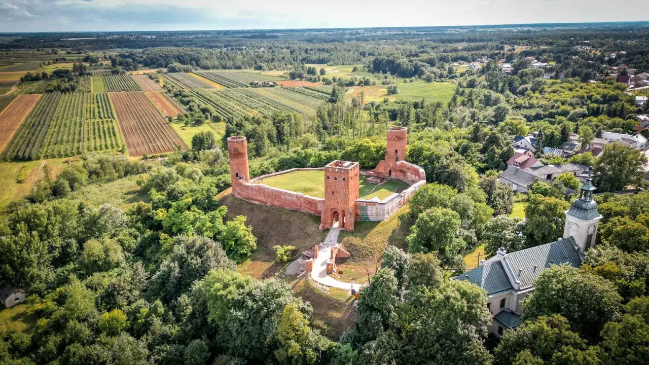 Ruiny zamku w otoczeniu zieleni, z widokiem na pola uprawne i kościół. To malowniczy zakątek, który mógłby znajdować się w największym województwie w Polsce.