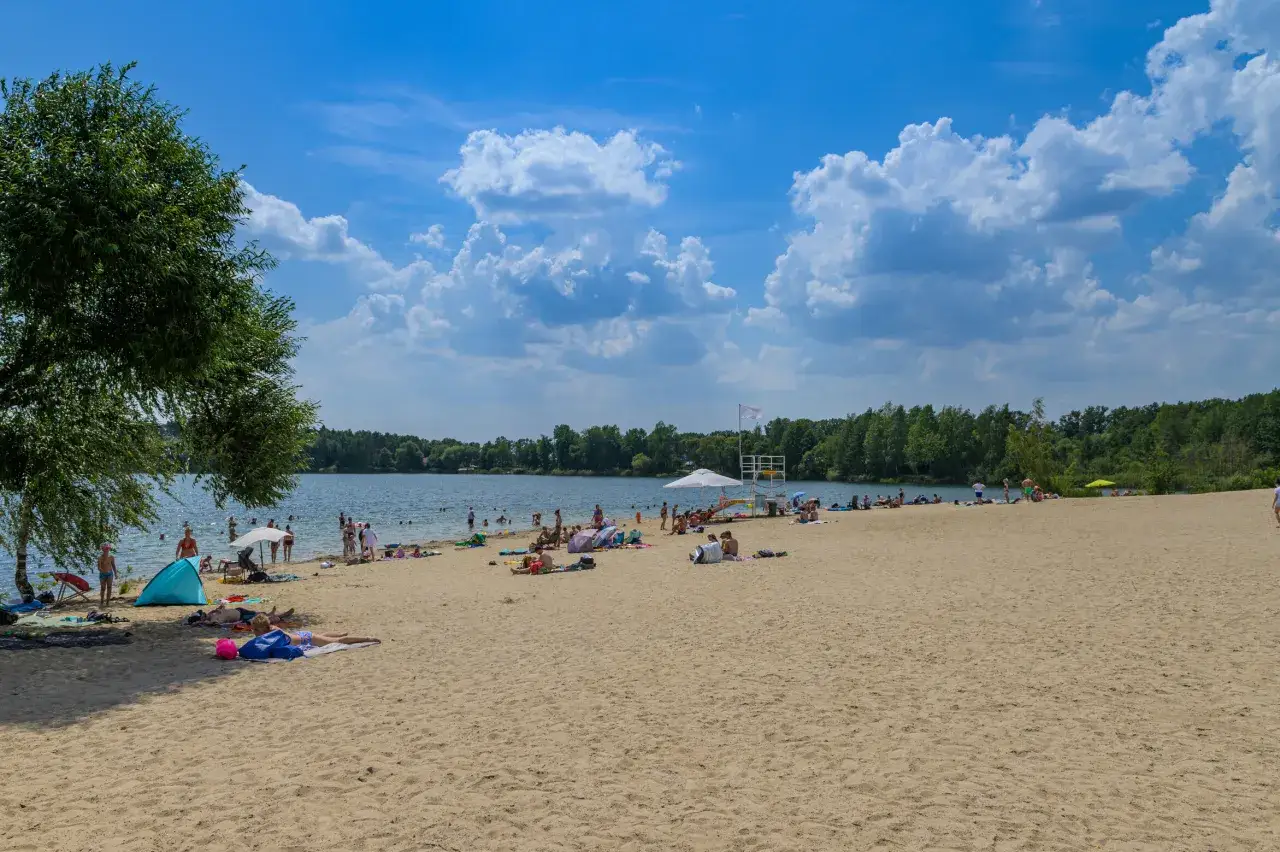 Słoneczny dzień nad jeziorem Głębocko Opolskie. Ludzie odpoczywają na piaszczystej plaży, kąpią się i opalają.