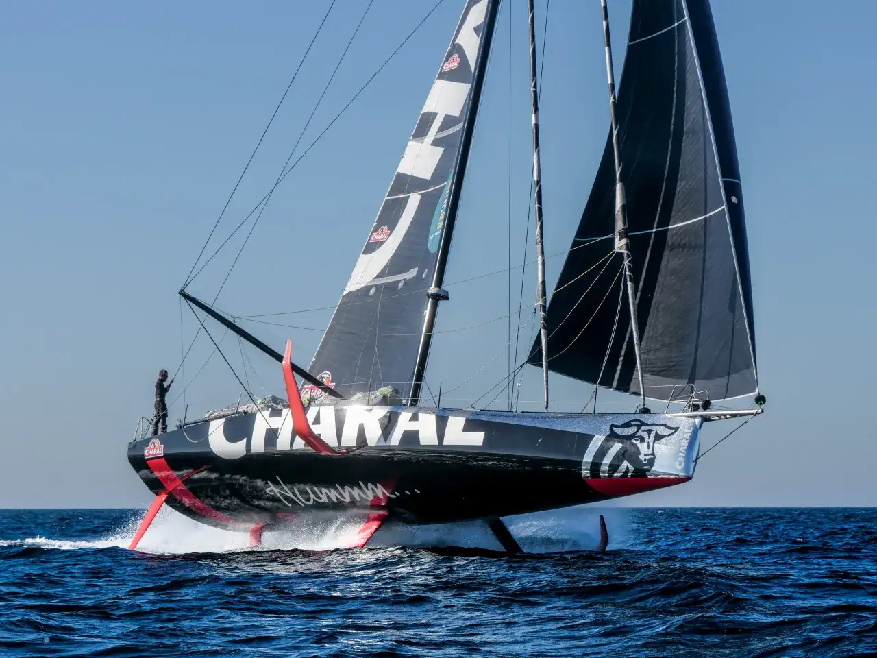 Un voilier IMOCA 60 pieds, le Charal, fend les vagues, ses foils rouges sortant de l'eau. Un marin veille sur le pont.