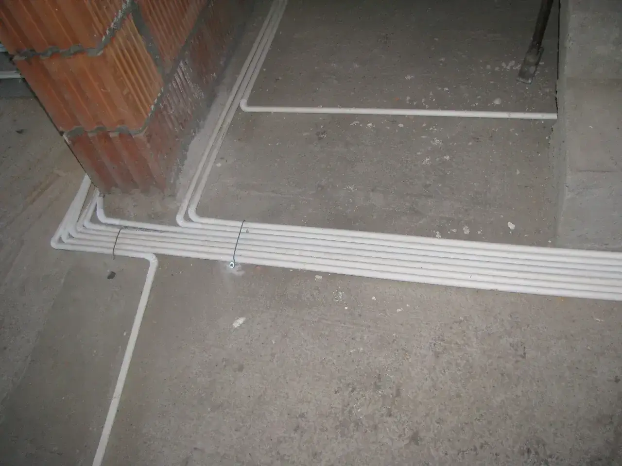 Przewody w posadzce bez peszla, ułożone równolegle obok ceglanego muru. Instalacja elektryczna w trakcie budowy.