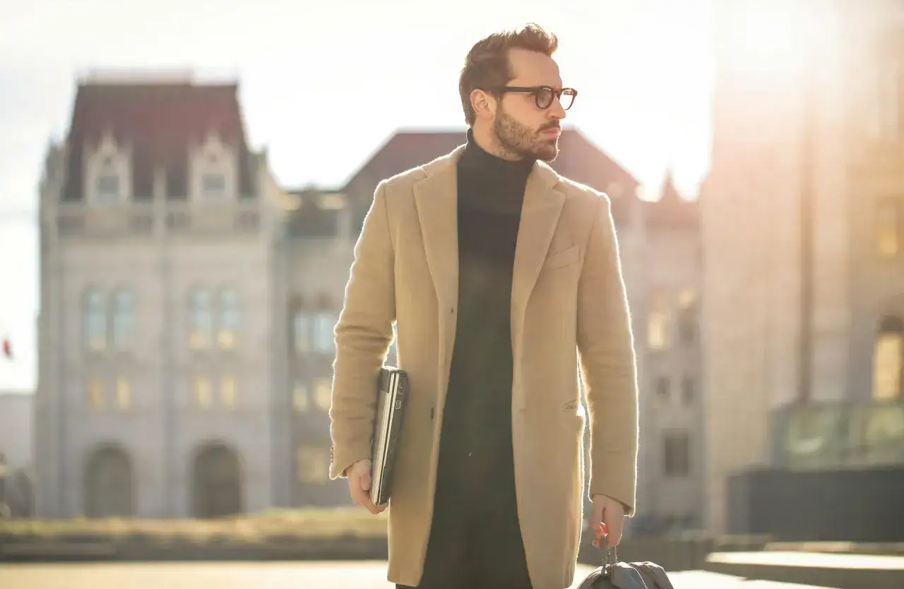 Mann im old money style mit gepflegten Herren Haaren, trägt Mantel und Brille, hält Laptop und Tasche.
