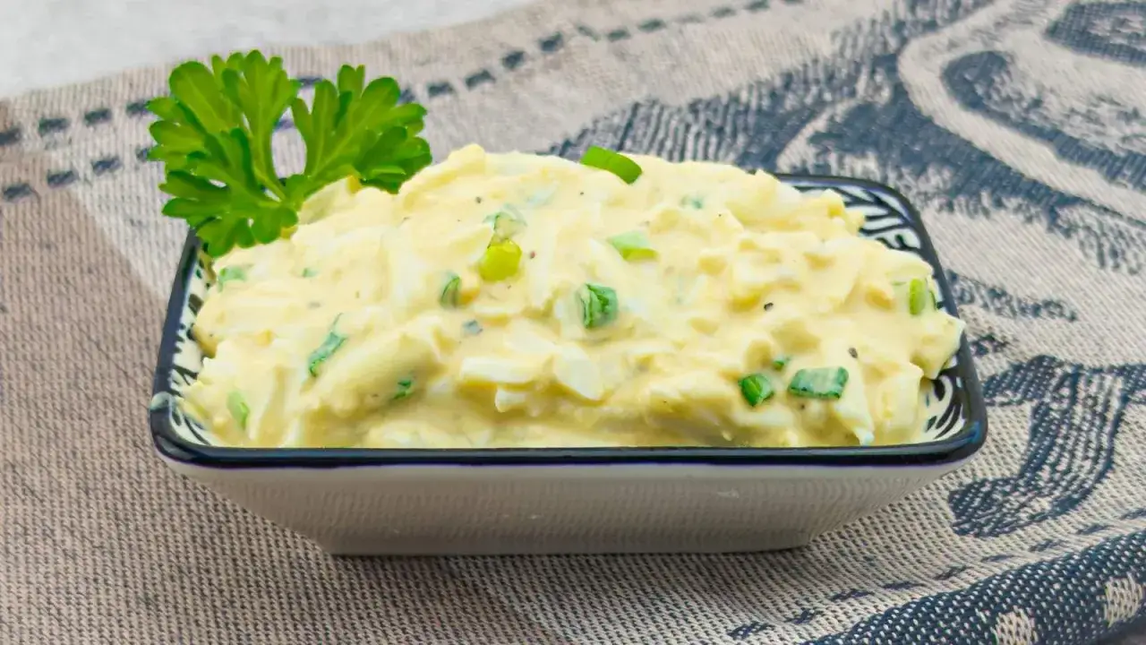 Pyszna pasta jajeczna z jogurtem, doprawiona szczypiorkiem i natką pietruszki, podana w miseczce na wzorzystym obrusie.