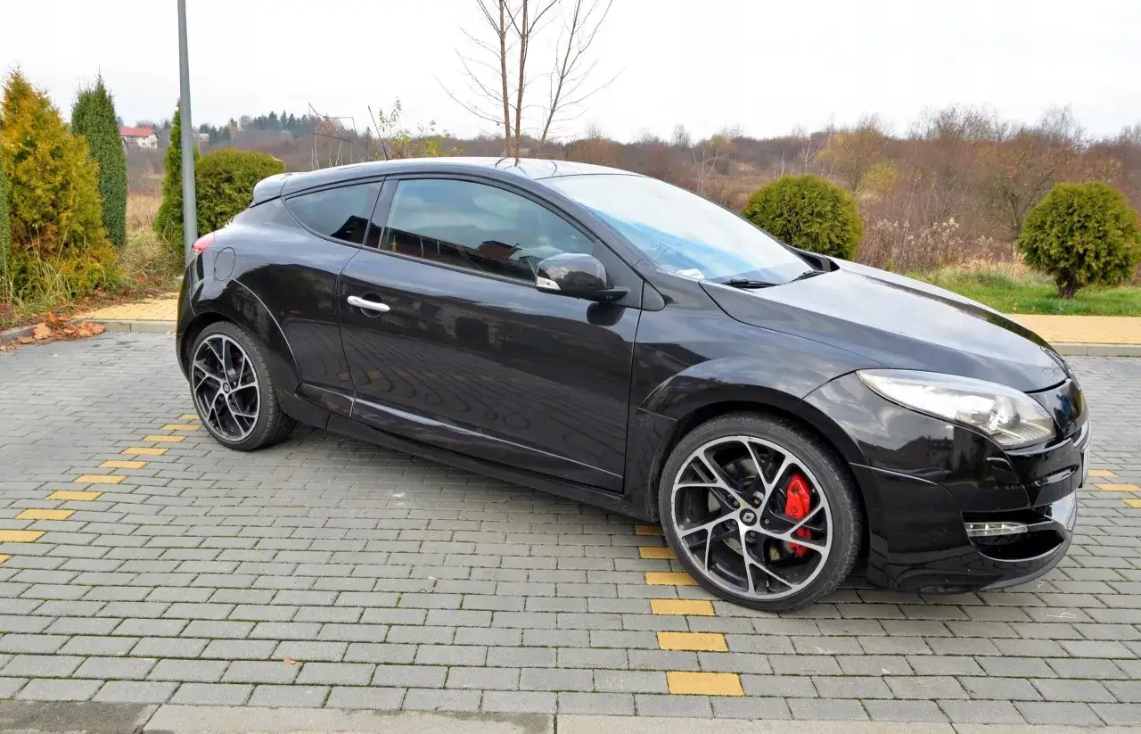 Czarny Renault Megane Coupe 3 z czerwonymi zaciskami hamulcowymi stoi na kostce brukowej, otoczony zielenią.