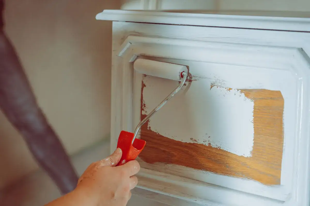 Eine Hand streicht mit einem kleinen Farbroller eine weiße Farbe auf ein Holzmöbelstück. Der Prozess des Holz streichen wird sichtbar.