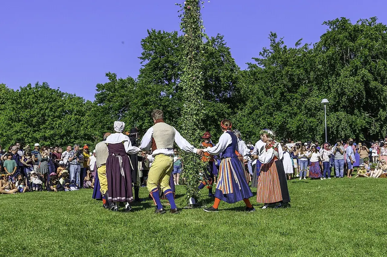 Midsommarhistoria: Människor i folkdräkter dansar runt en midsommarstång på en grön gräsmatta, omgivna av en publik.