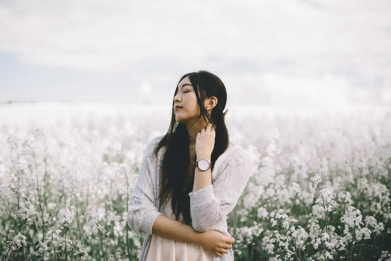 A young woman with eyes closed, touching her neck, stands in a field of white flowers. The blurry background emphasizes her peaceful moment.
