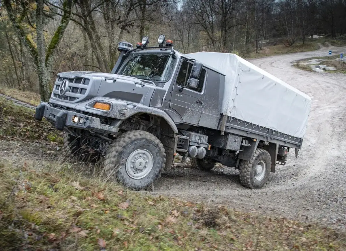 Szary Mercedes Zetros z plandeką pokonuje wyboisty teren leśny.