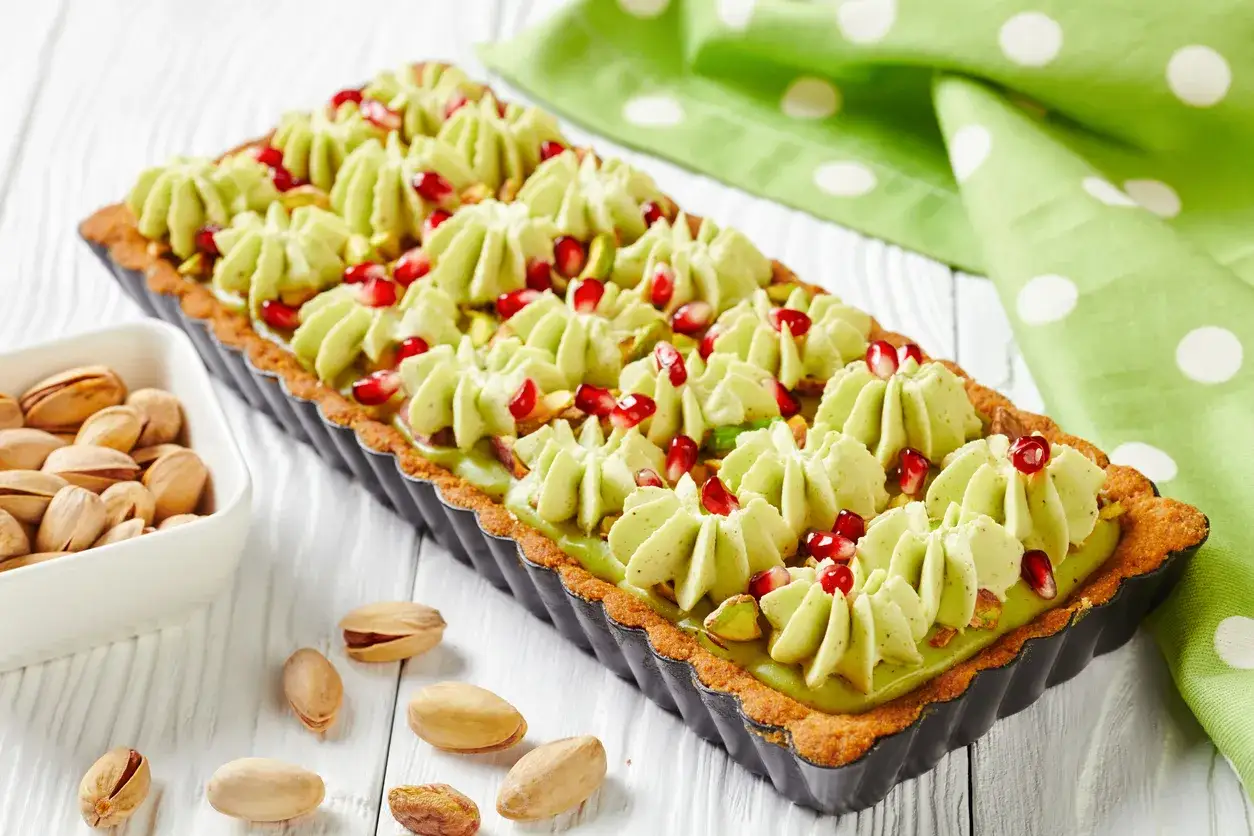 Pyszny mazurek pistacjowy z malinami, udekorowany kremem pistacjowym i granatem. Obok miseczka pistacji.