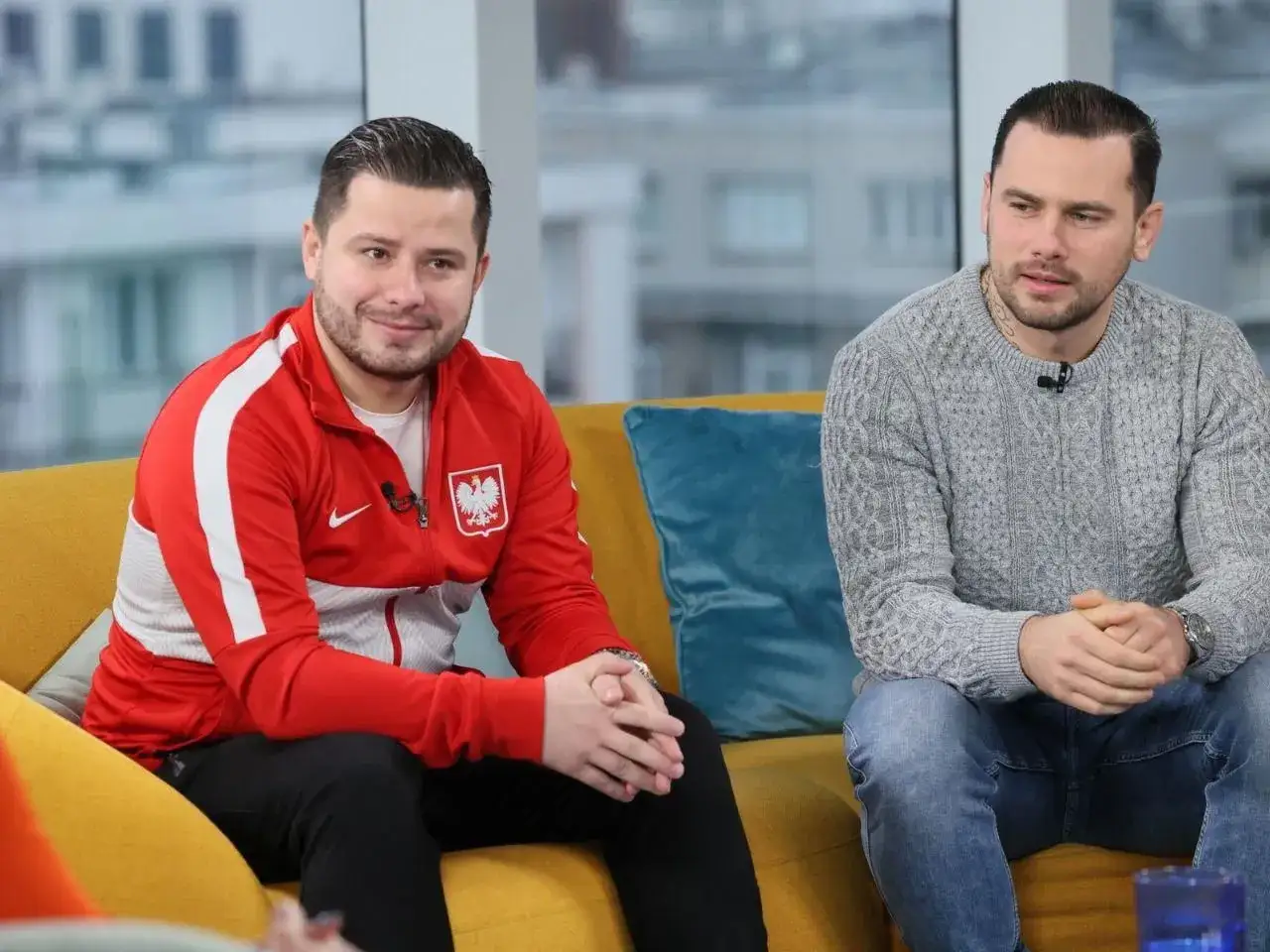 Grzegorz Collins, w wieku około 30 lat, w czerwonej bluzie z orłem, siedzi obok kolegi w szarym swetrze.