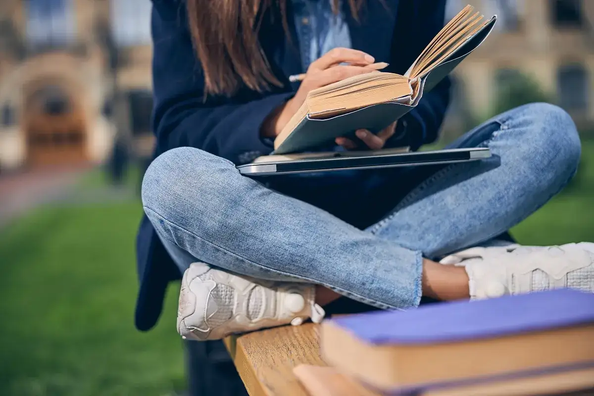 Studentka w jeansach i marynarce siedzi na ławce, czyta książkę i notuje. Studium daje jej wiedzę i rozwój.