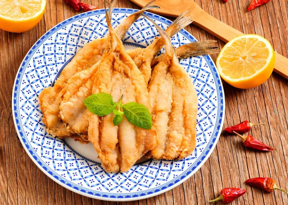 Un plato de boquerones fritos dorados, decorado con menta, junto a limones y guindillas rojas sobre una mesa de madera.