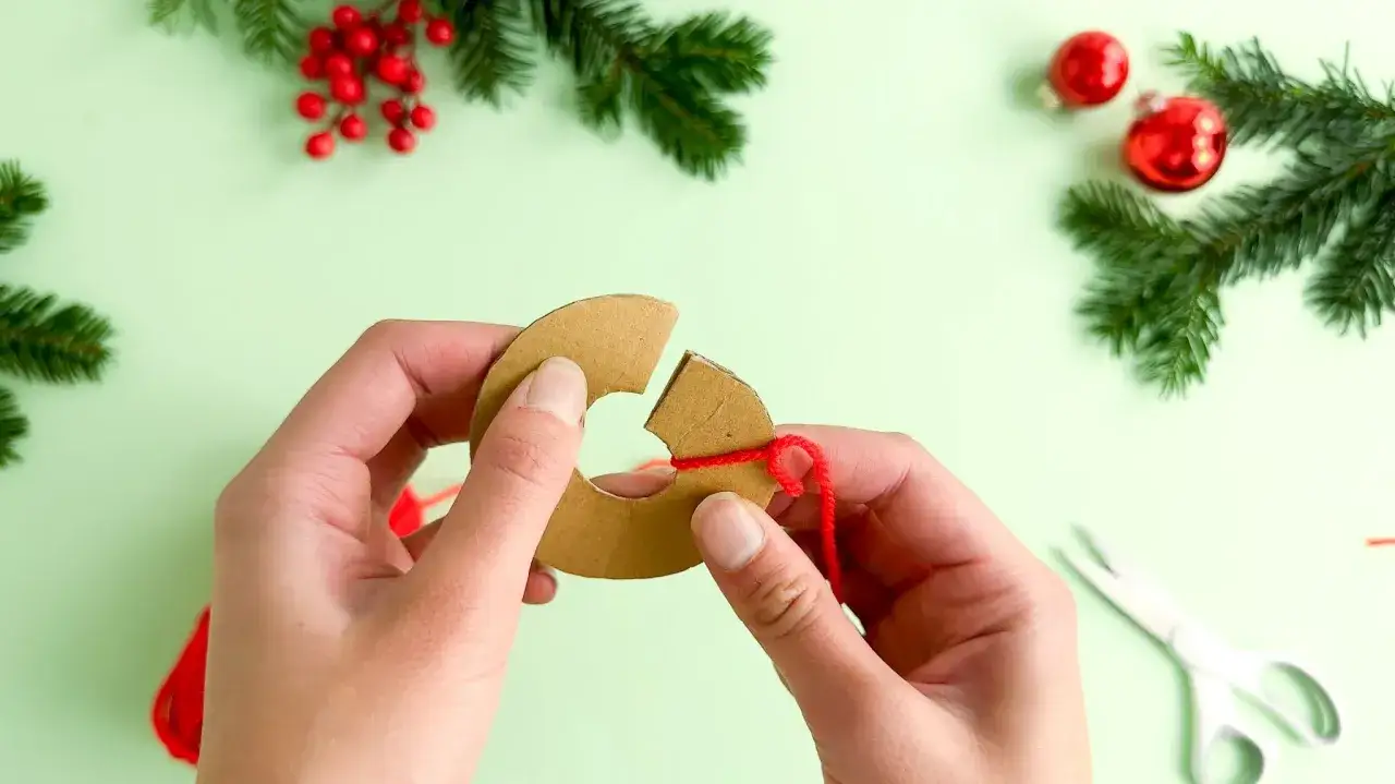 Hände wickeln roten Faden um Pappring für **Basteln mit Pompons Weihnachten**. Tannenzweige und Kugeln im Hintergrund.