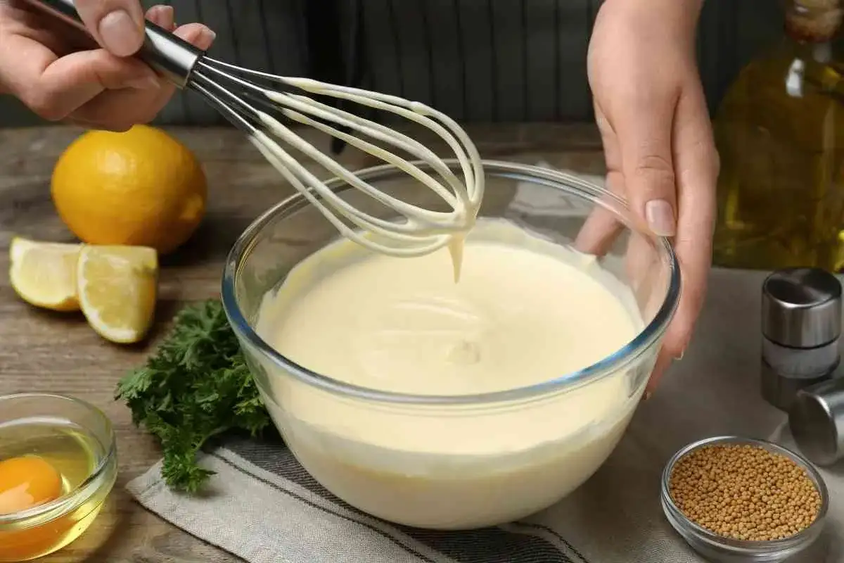 Manos batiendo mayonesa casera en un bol de cristal. Ingredientes como lim&oacute;n, huevo y perejil alrededor.