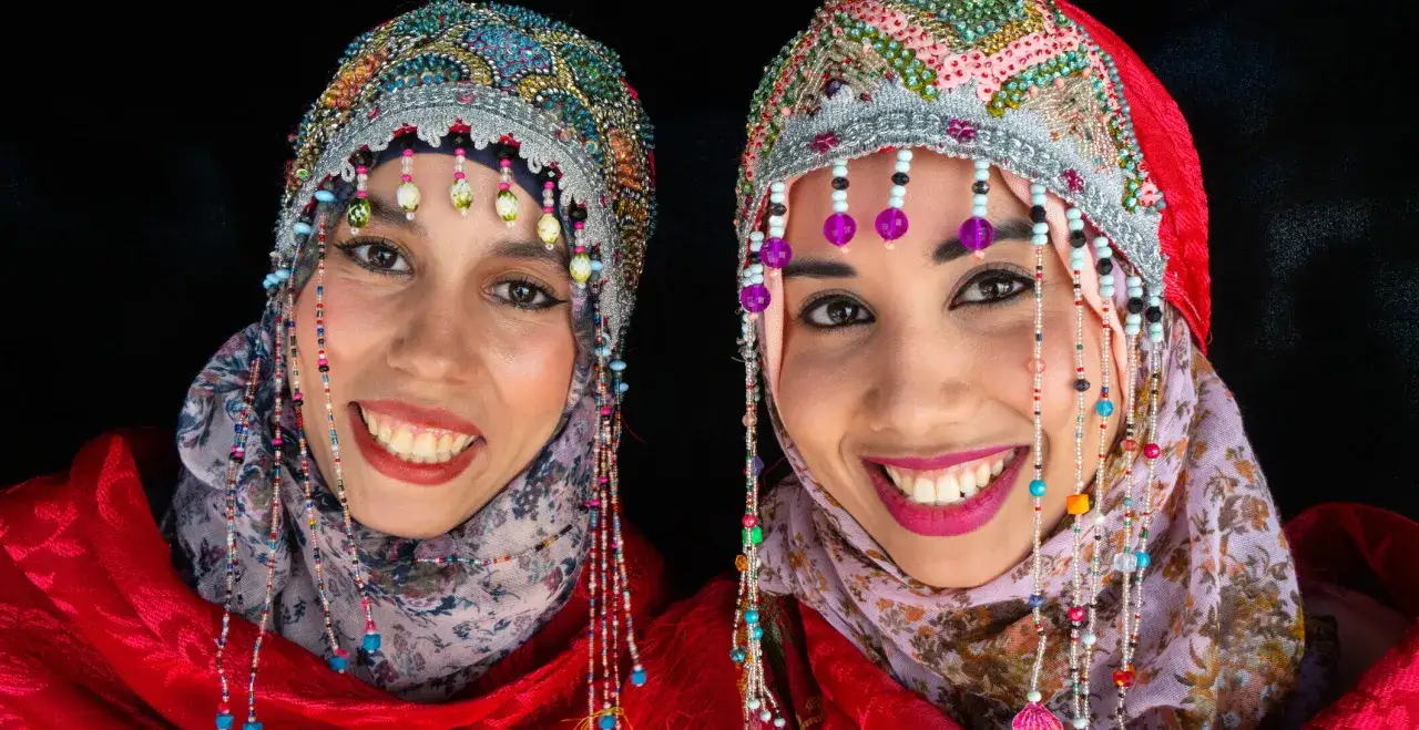 Zwei l&auml;chelnde Frauen mit reich verzierten Kopfbedeckungen und Perlenketten, die an traditionelle Berber-Kleidung erinnern.