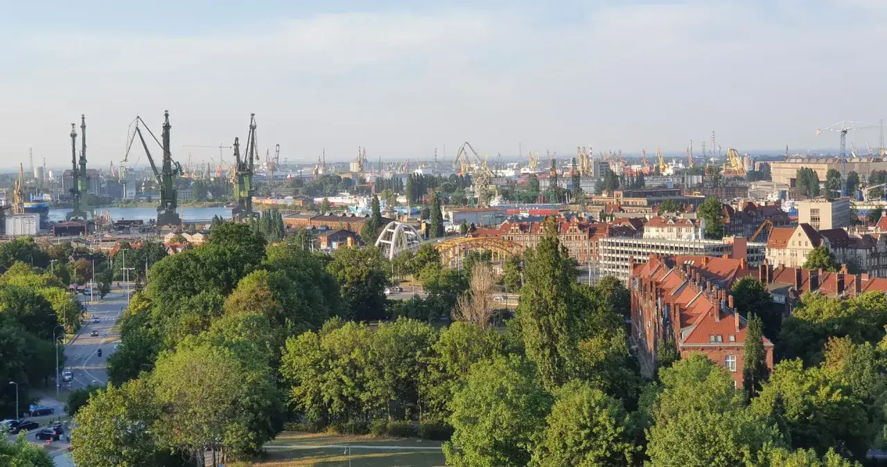 Widok na port z dźwigami i miasto z czerwonymi dachami, otoczone bujną zielenią. Długie ogrody miejskie przechodzą w przemysłowy krajobraz.