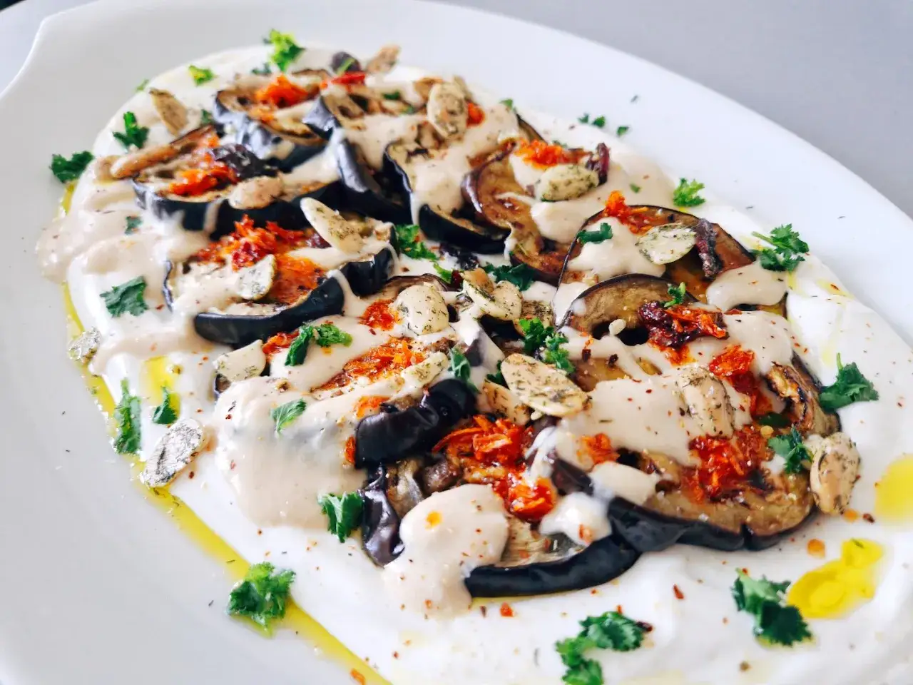 Hummus de berenjena cremoso, cubierto con rodajas de berenjena asada, hierbas frescas y un chorrito de aceite de oliva.