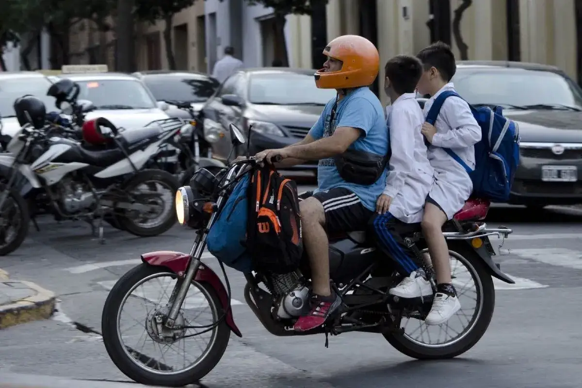 Un hombre lleva a dos niños en moto, uno adelante y otro atrás, todos con mochilas.