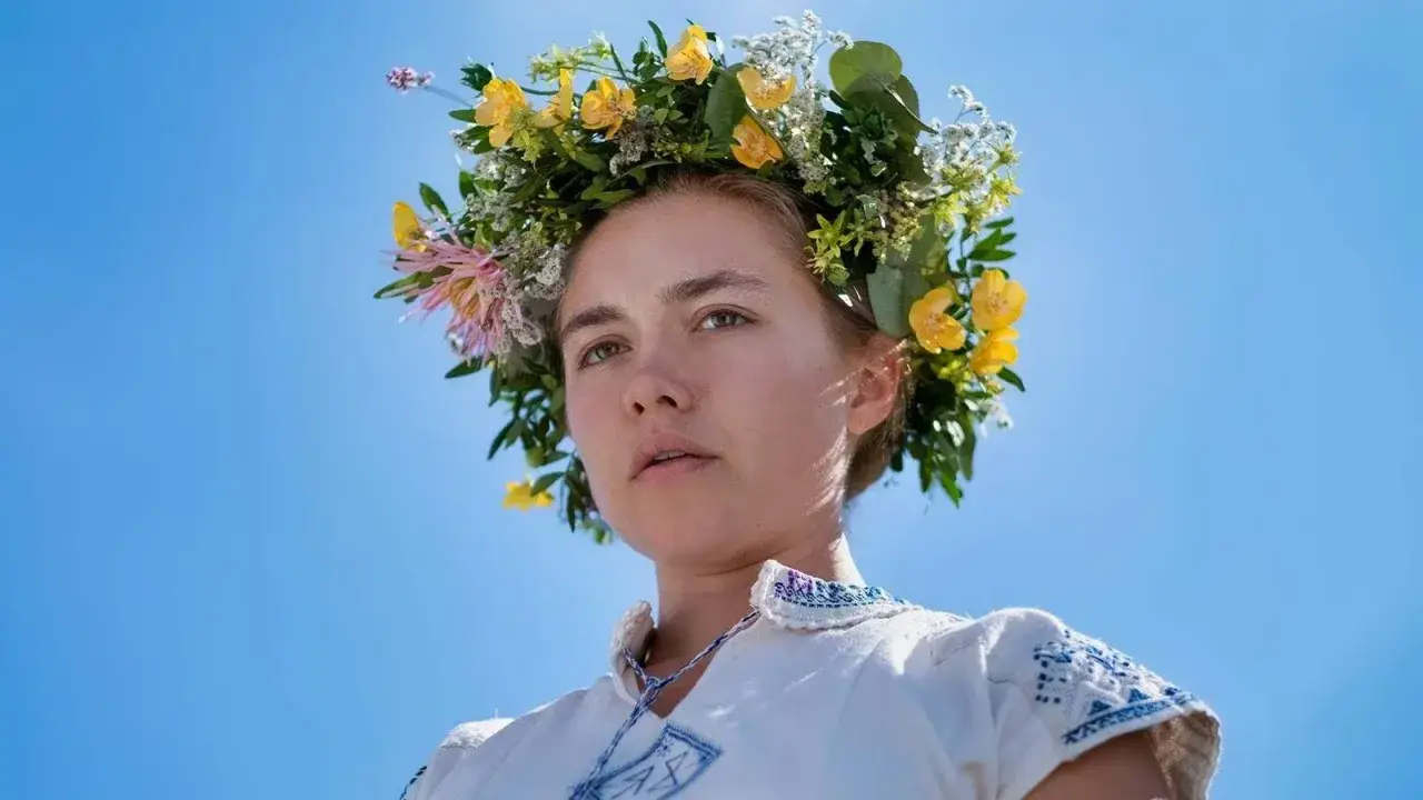 En kvinna med blomsterkrans i h&aring;ret, kl&auml;dd i en broderad vit kl&auml;nning, en scen fr&aring;n filmen Midsommar mot en klarbl&aring; himmel.