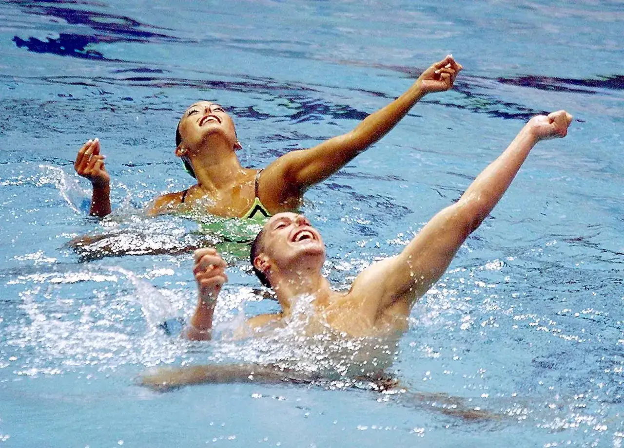 Dwóch zawodników, mężczyzna i kobieta, wykonuje choreografię w wodzie. Ich ramiona są uniesione, a twarze skierowane ku górze z wyrazem radości. To dynamiczne pływanie synchroniczne mężczyzn.