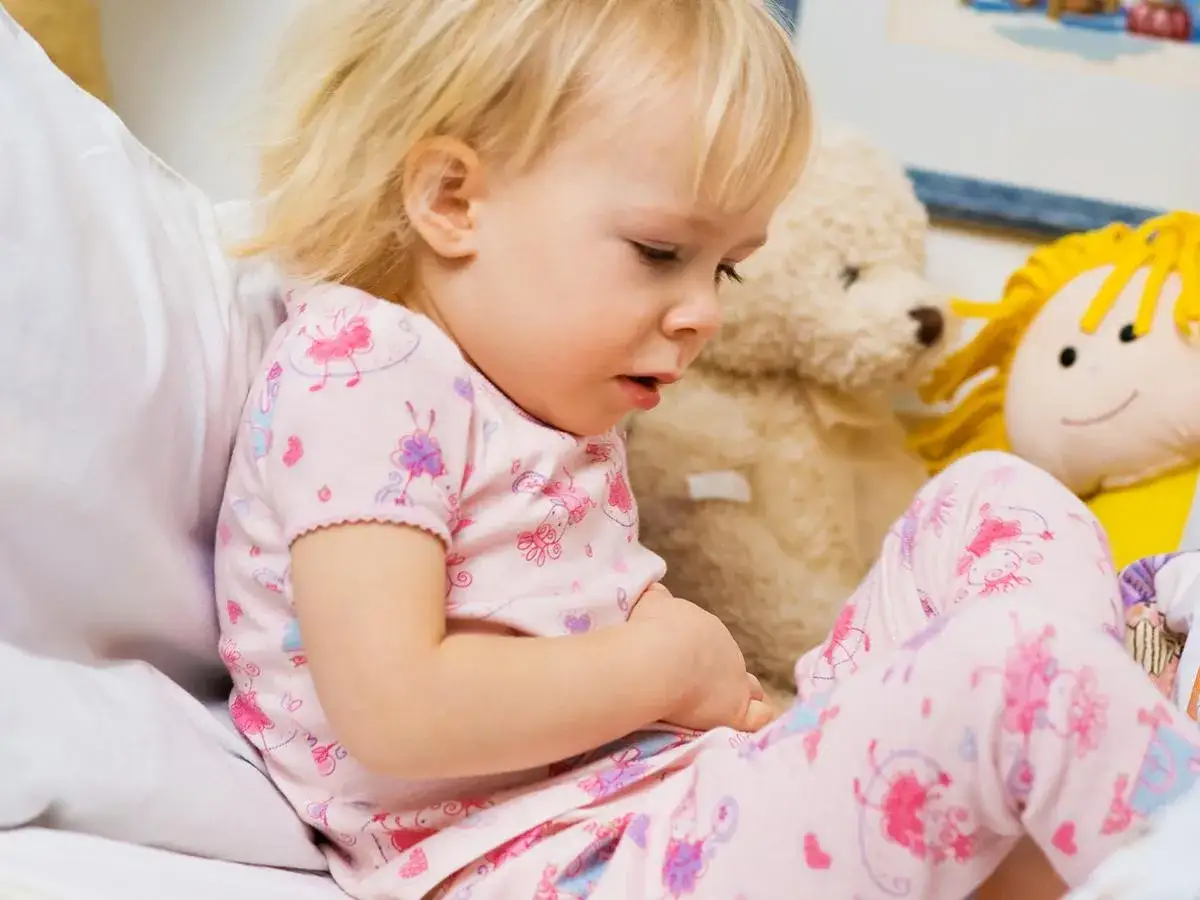 Bambina con pigiamino rosa, che si tiene la pancia, forse per vomito e diarrea. Orsacchiotto e bambola sullo sfondo.