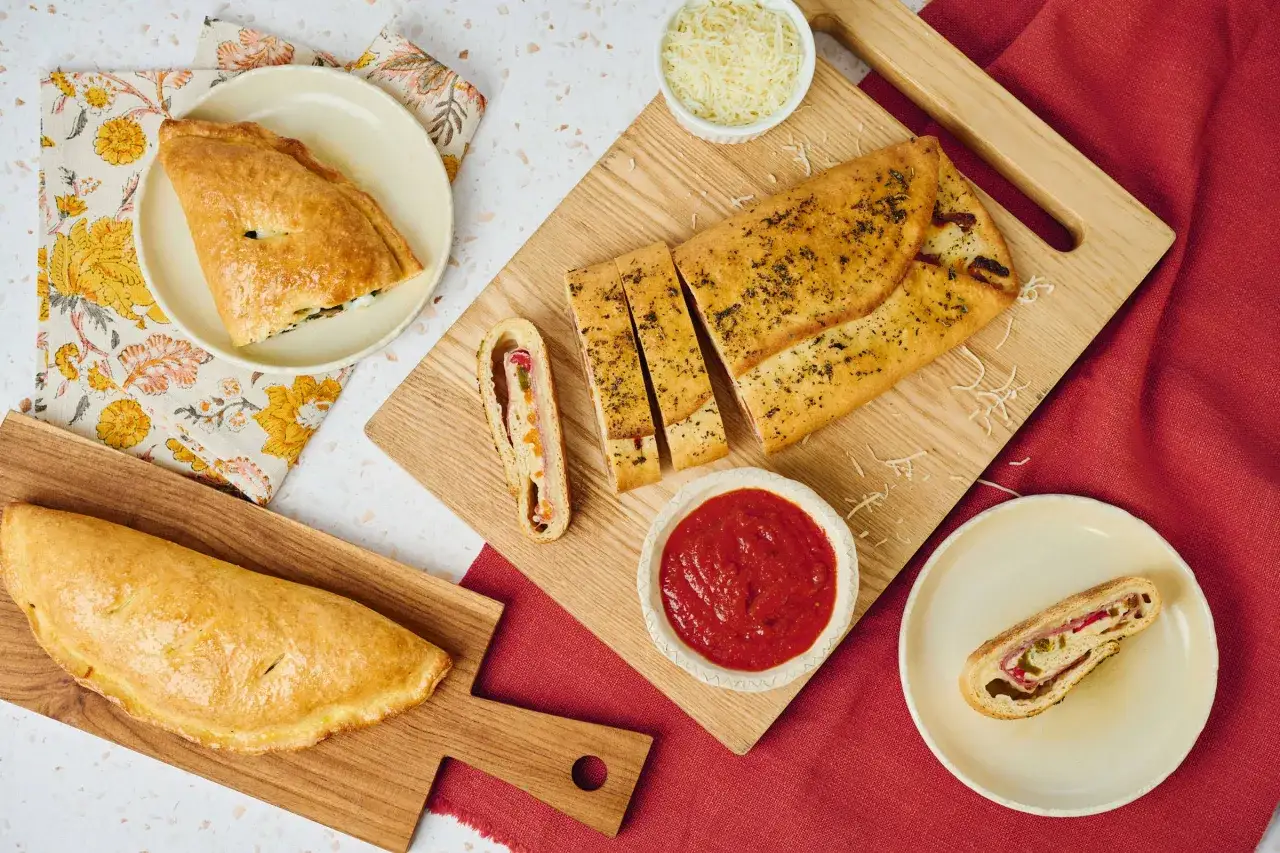 Två gyllene calzoner, en hel och en skivad, med fyllning av kött och grönsaker. Serveras med tomatsås och riven ost. Perfekt för att njuta av utan att oroa sig för calzone kalorier.