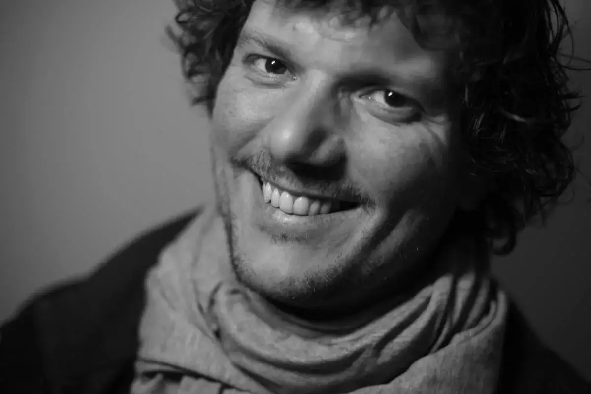 Uśmiechnięty Filip Ćwik w szarym szaliku, z burzą kręconych włos&oacute;w i charakterystycznym wąsikiem.