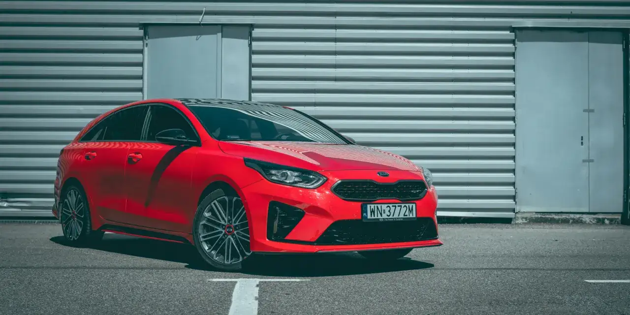 Czerwony Kia Proceed GT z czarnym dachem zaparkowany przed szarą, falistą ścianą.