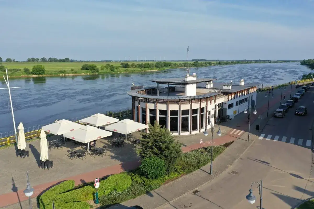 Tczew atrakcje: nowoczesny budynek nad Wisłą z tarasem i ogr&oacute;dkiem letnim, otoczony zielenią i wodą.
