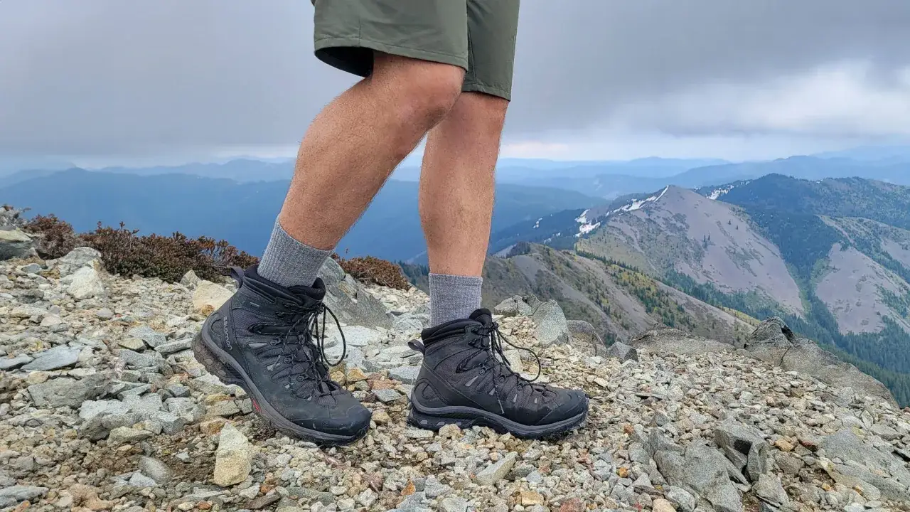 A hiker's legs in hiking fit boots and grey socks stand on a rocky mountain summit, with a vast, layered mountain range stretching into the distance under a cloudy sky.