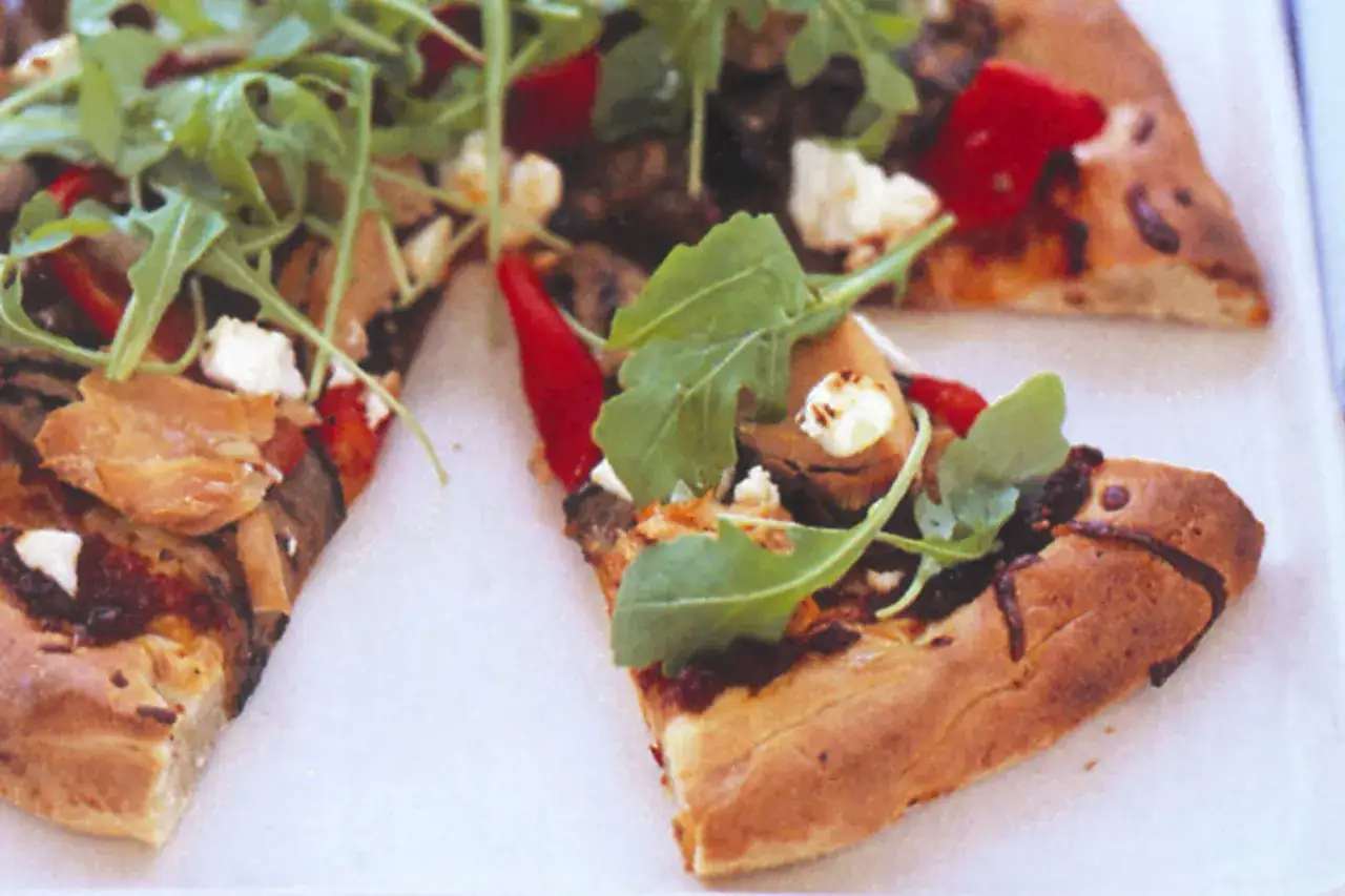Tranches de pizza au thon garnies de roquette fra&icirc;che, de poivrons rouges et de fromage &eacute;miett&eacute;, sur une cro&ucirc;te dor&eacute;e.