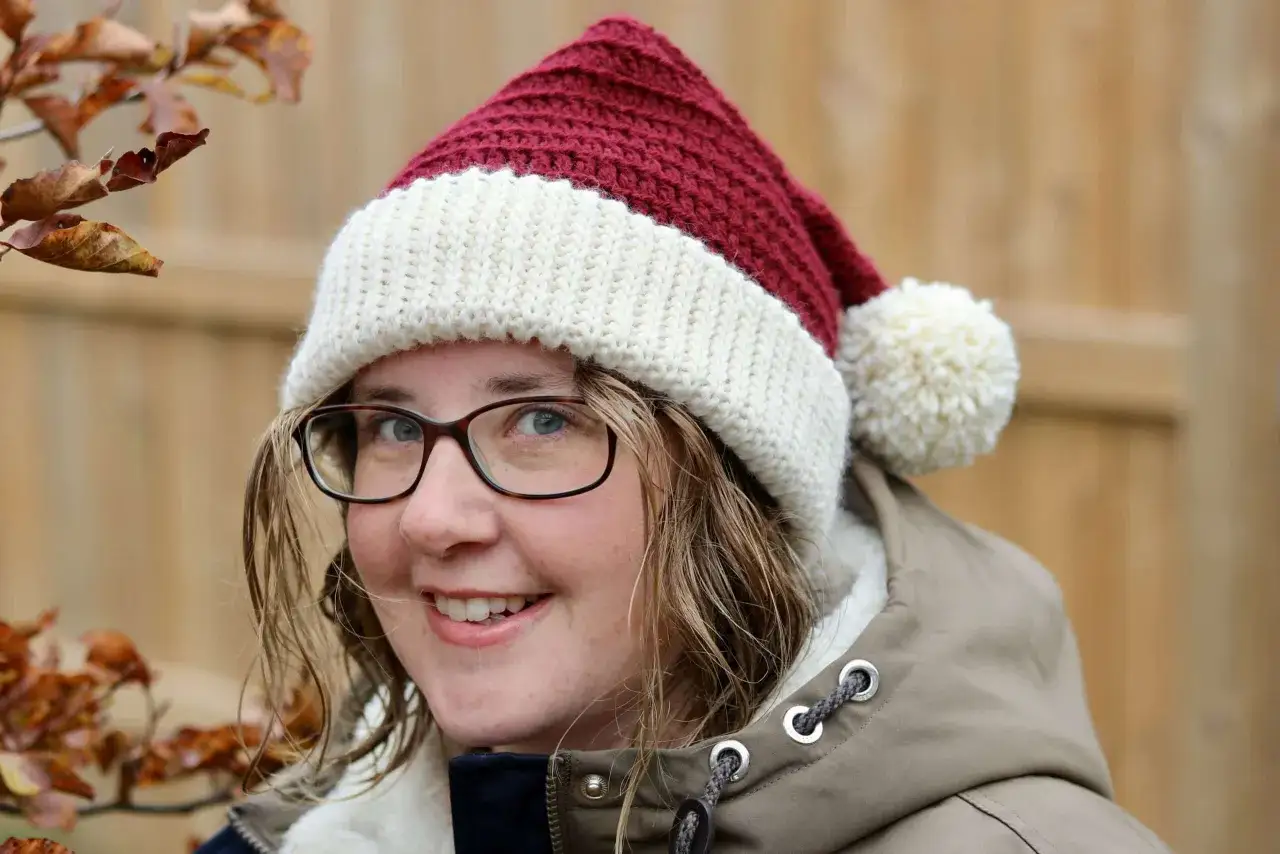 En glad person med glasögon bär en röd och vit virkad tomteluva med en fluffig pompom.
