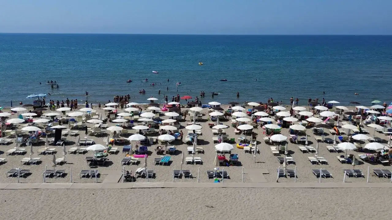 Spiaggia affollata a Paestum mare, con ombrelloni bianchi e lettini sulla sabbia dorata. Persone nuotano e si rilassano in un'atmosfera estiva.