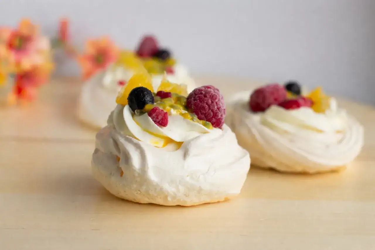 Mini bezy pavlova z bitą śmietaną i owocami: malinami, borówkami, pomarańczą i marakują.