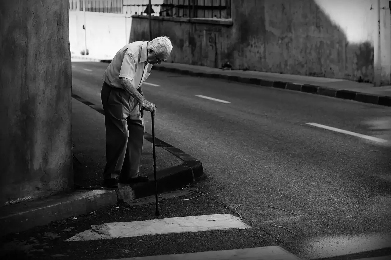 Une photo dans la rue : un vieil homme se penche sur sa canne, le regard perdu, au bord d'une route déserte.